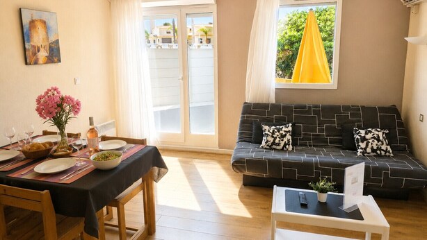 A dining area with a table set for two, a sofa, and a small side table with a remote control.