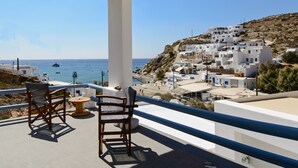 A balcony with two wooden chairs and a small round table, overlooking a coastal village with white buildings and boats on the sea.