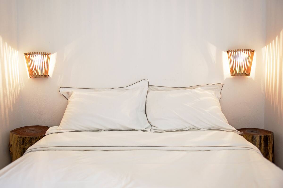 A neatly made bed with white linens, two bedside tables with wooden tops, and wall-mounted lamps on either side.
