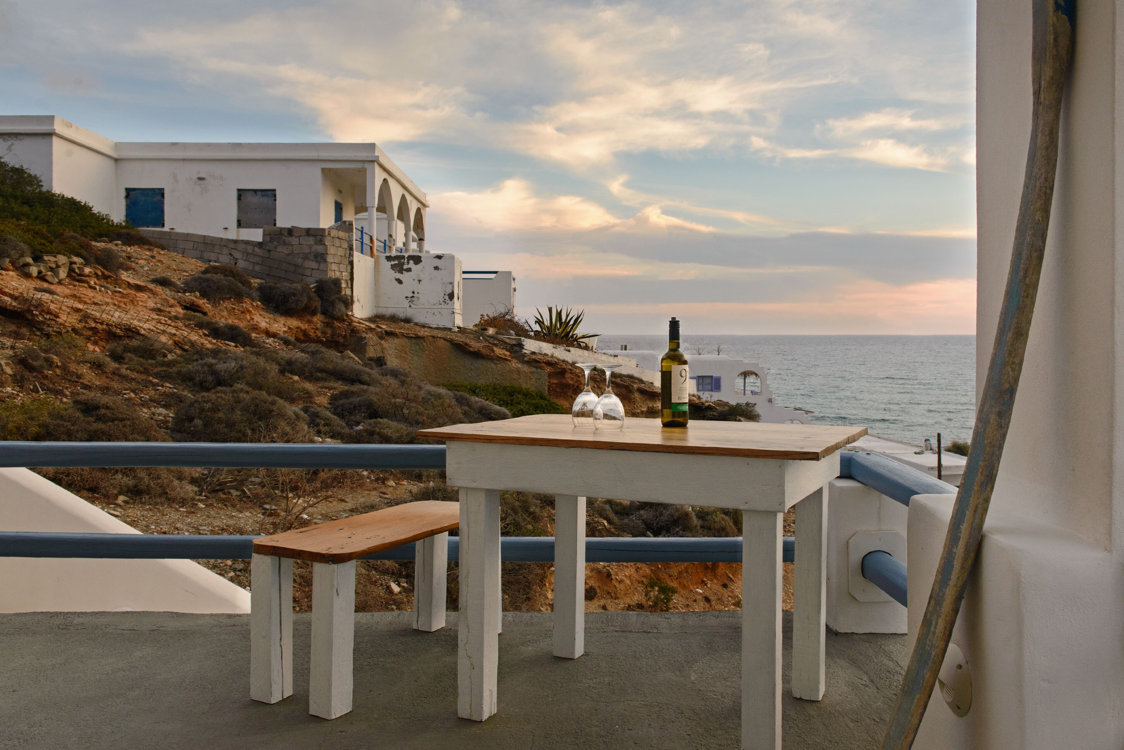 A seaside terrace with a table and bench, overlooking the sea and a white building with arches.