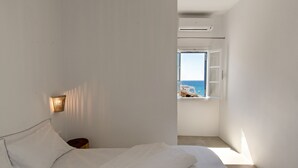A bedroom with a bed, a bedside table, a wall-mounted light, and a window with a view of the sea.