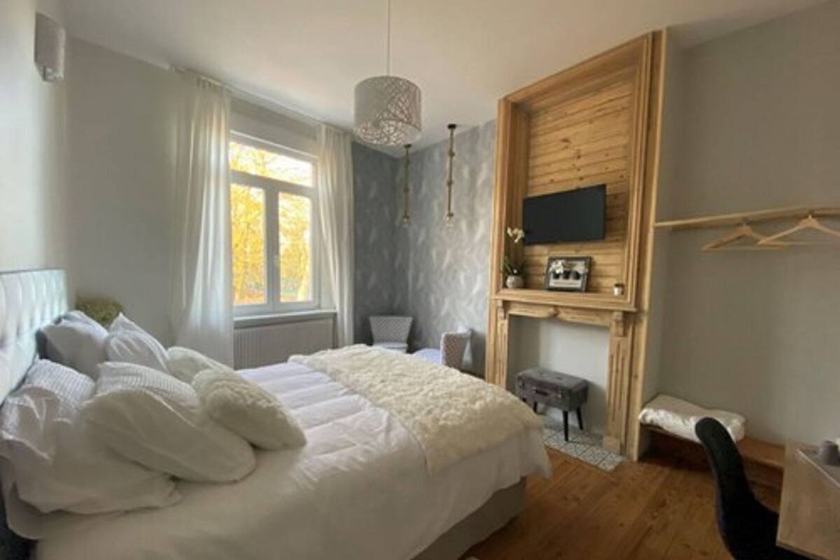 A bedroom with a large bed, a wooden wall panel, a TV mounted above a fireplace, and a window with curtains.