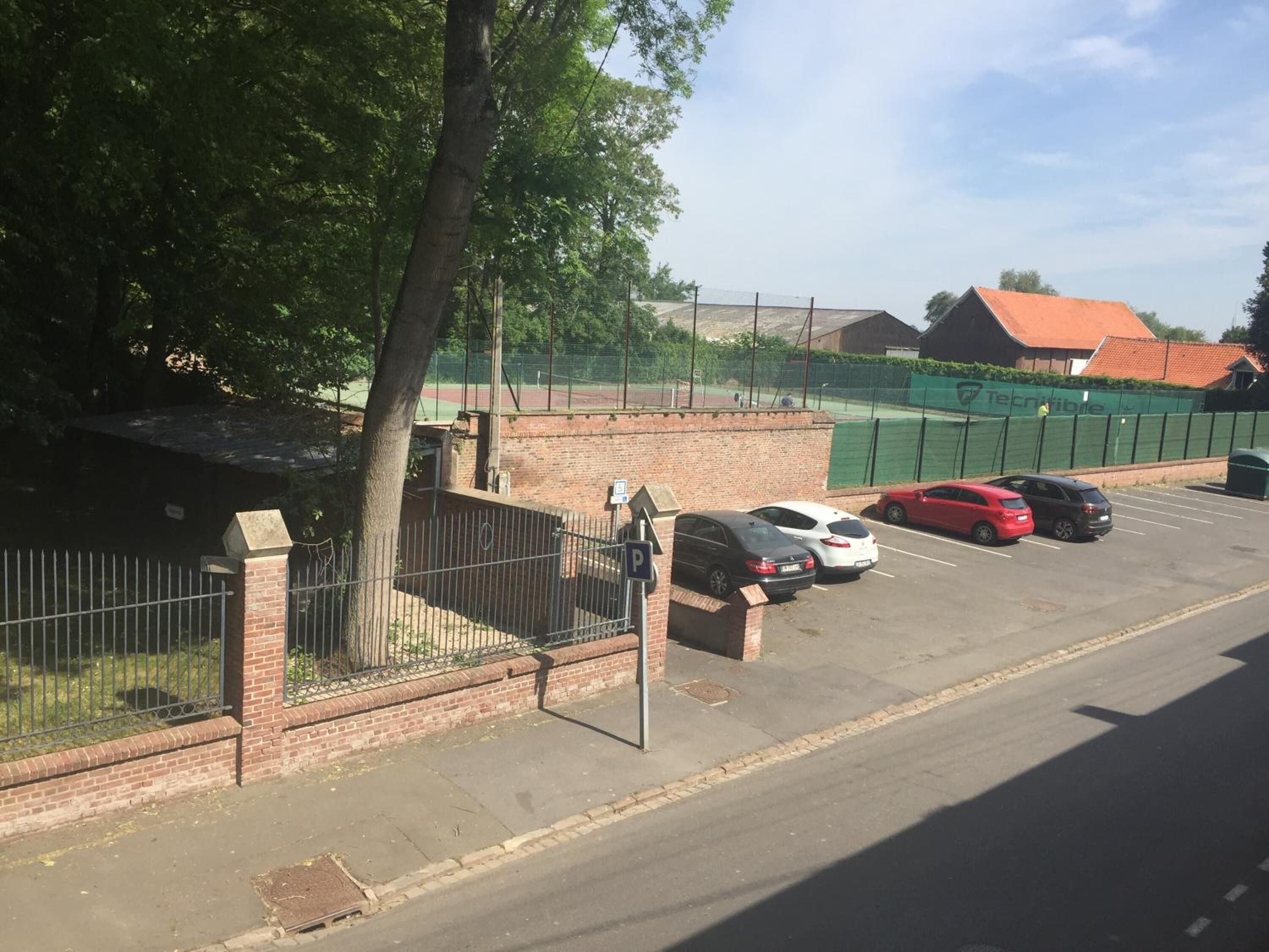 Een parkeerplaats met verschillende auto's, een bakstenen muur en een tennisbaan op de achtergrond.