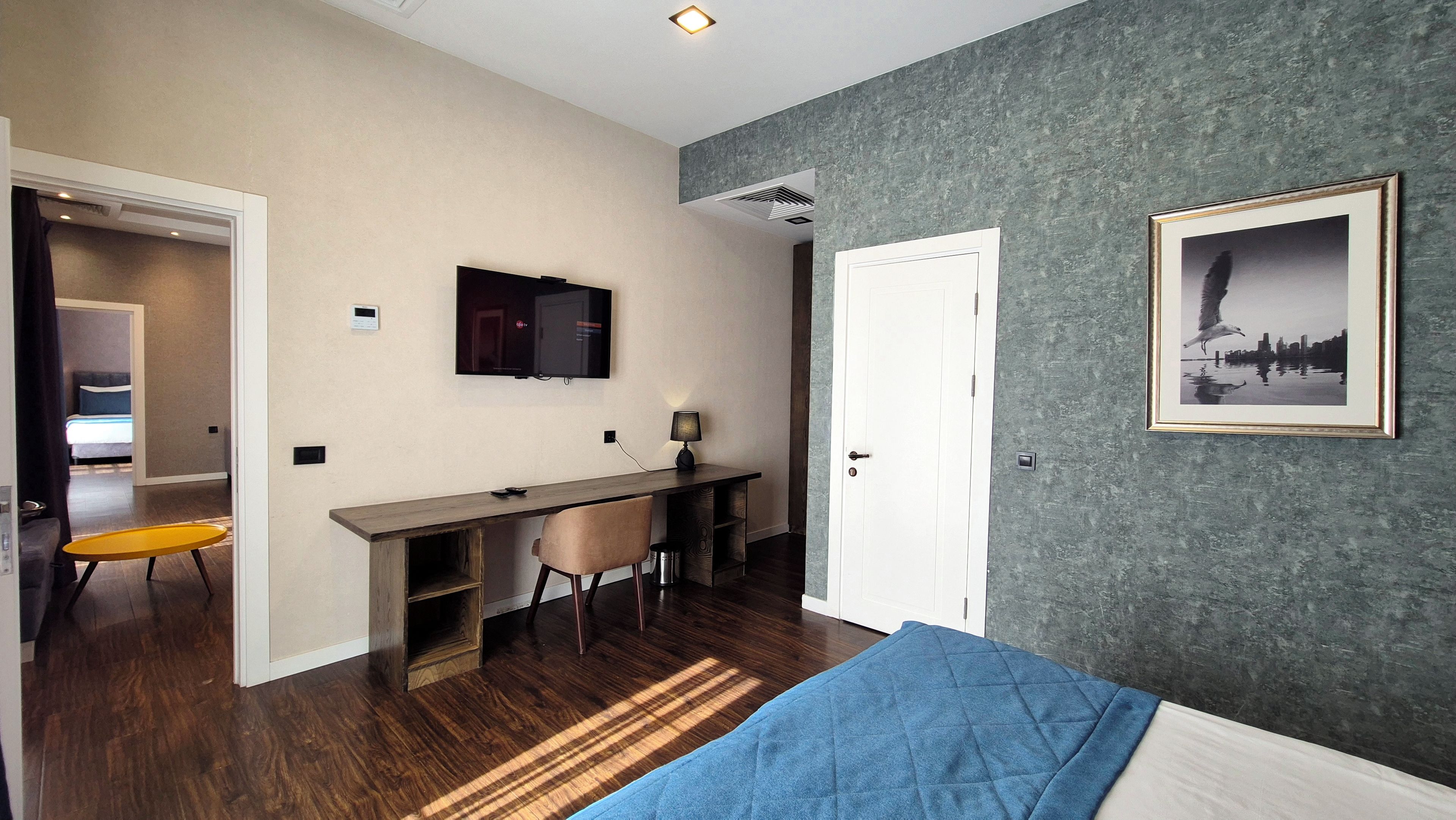 A bedroom with a bed, a desk, and a chair. There is a door, a wall-mounted TV, and a framed picture.