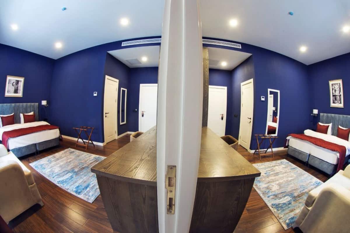Una habitación de hotel con una gran mesa de madera, dos camas con ropa de cama roja y blanca, y paredes azules.