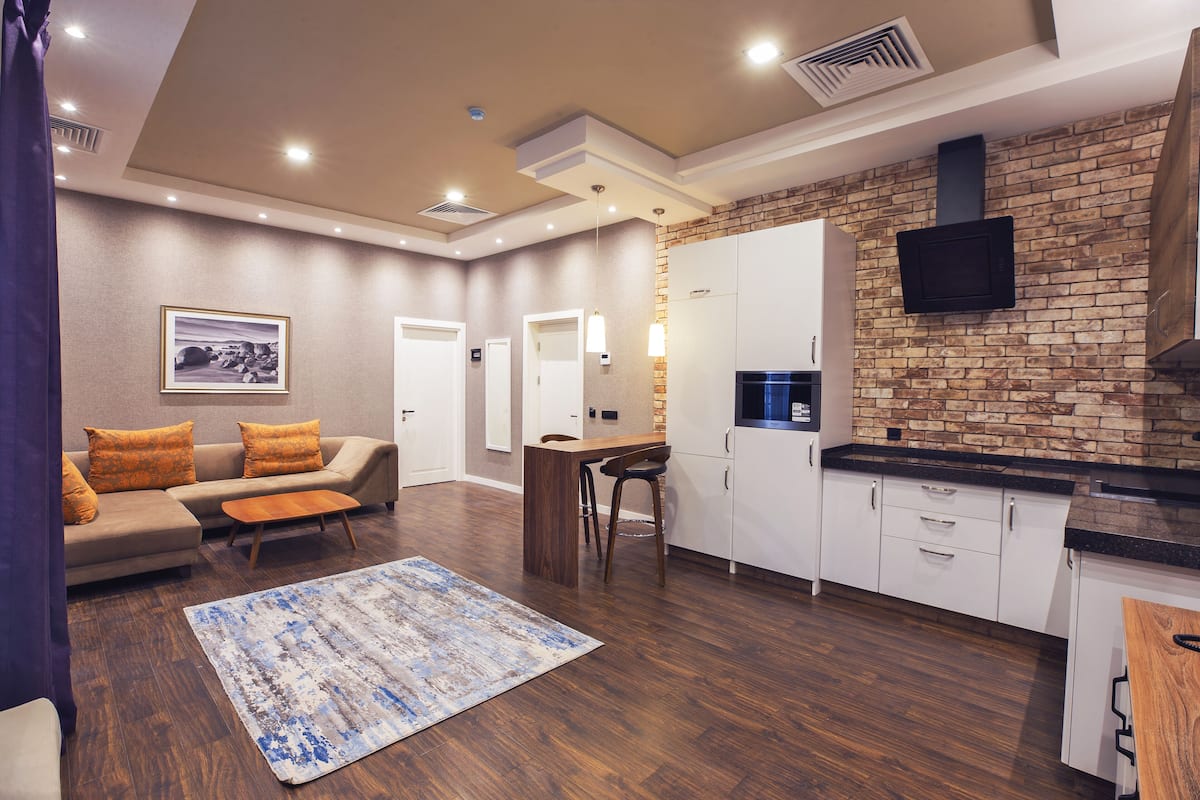 Una sala moderna con sofá, mesa de centro y alfombra. Se observa una zona de cocina con pared de ladrillo, armarios y televisor.