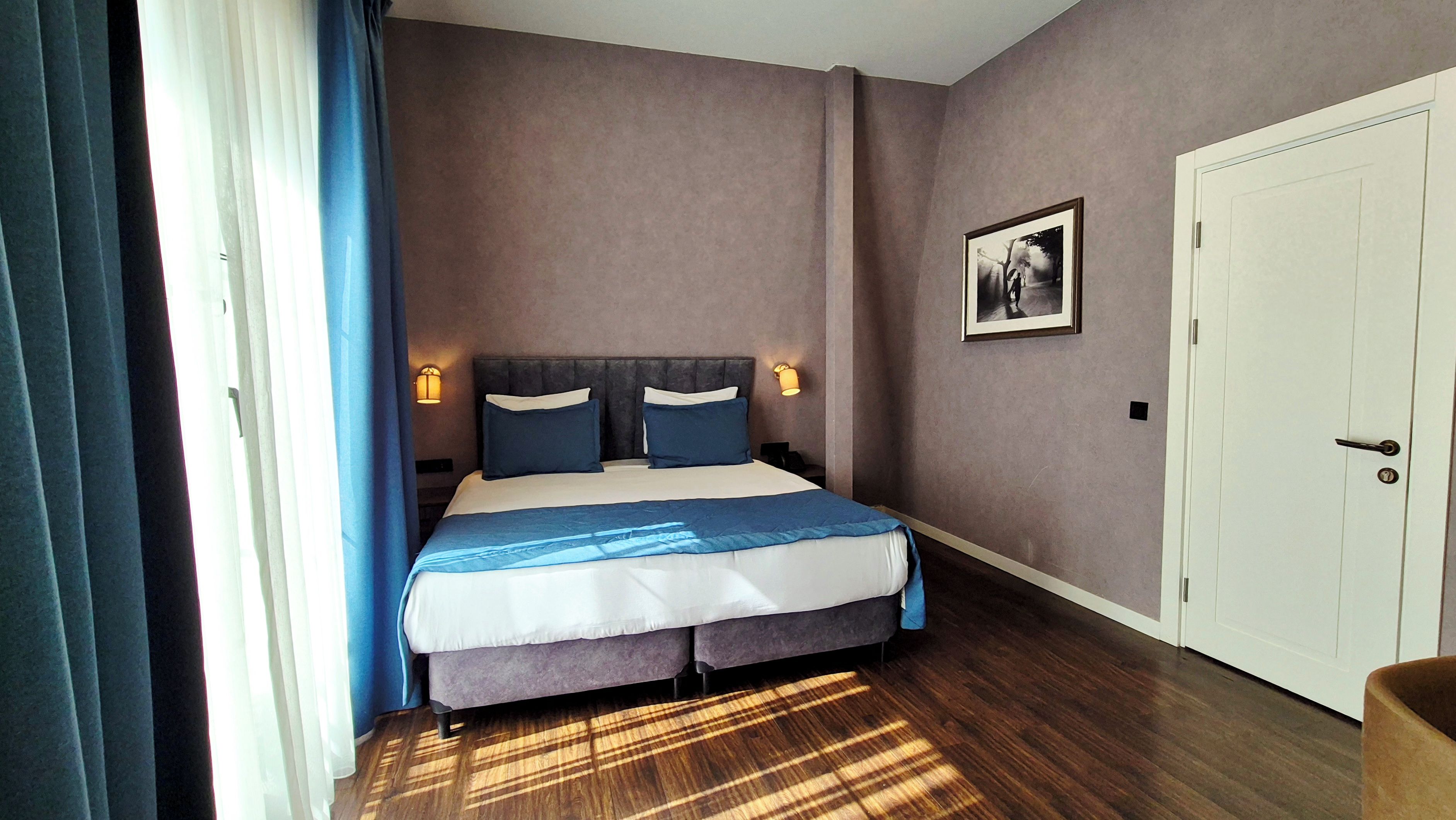 A hotel room with a bed, bedside lamps, a door, and a framed picture on the wall.
