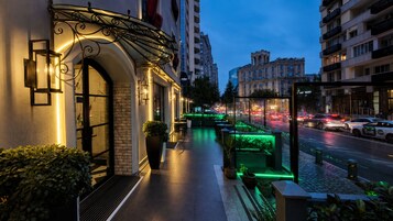 Entrada de hotel com fachada de tijolos, área de estar ao ar livre e vista para a rua com carros.