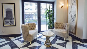 A lobby with two plush armchairs, a marble-patterned floor, a potted plant, and a large window with a view of buildings.