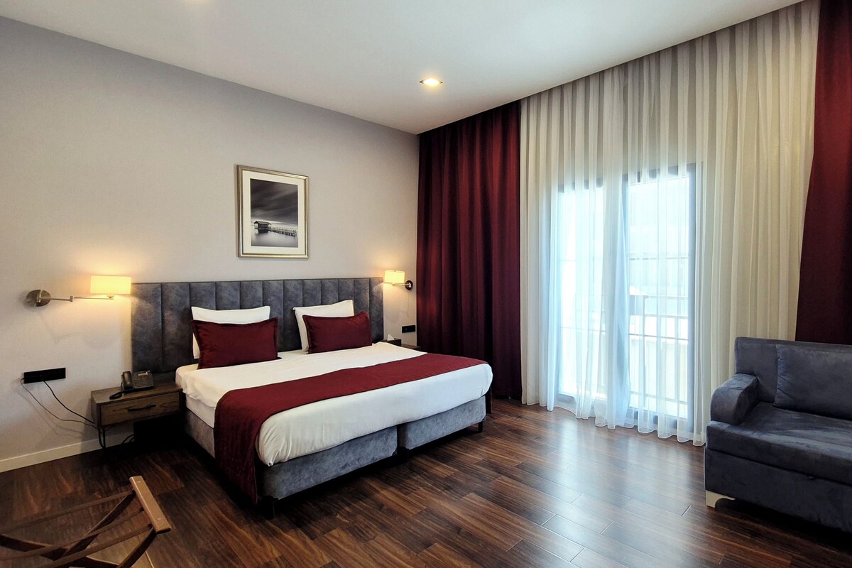 A hotel room with a bed, a nightstand, a lamp, a sofa, and a window with curtains.