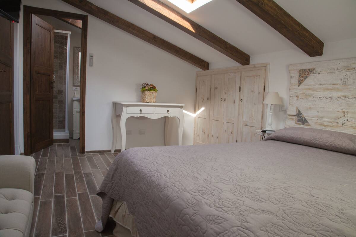 Une chambre avec un grand lit, des poutres en bois au plafond, une commode blanche et une porte donnant sur une autre pièce.