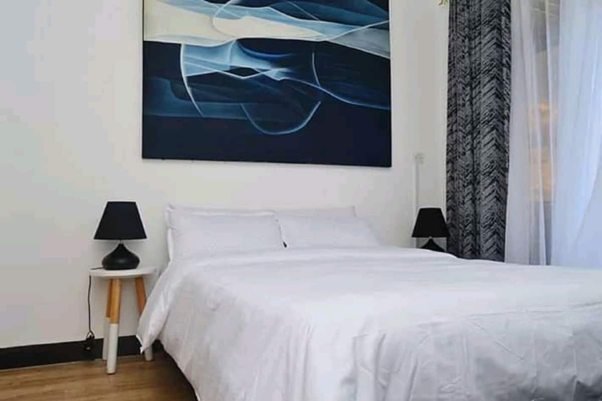 Une chambre à coucher avec un lit, deux tables de chevet, un tableau abstrait accroché au mur et des rideaux en voile.