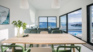 A modern dining area with a wooden table and green chairs, large windows with a view of the sea, and a sofa with cushions. - The Olive Exclusive holiday home (Okrug)