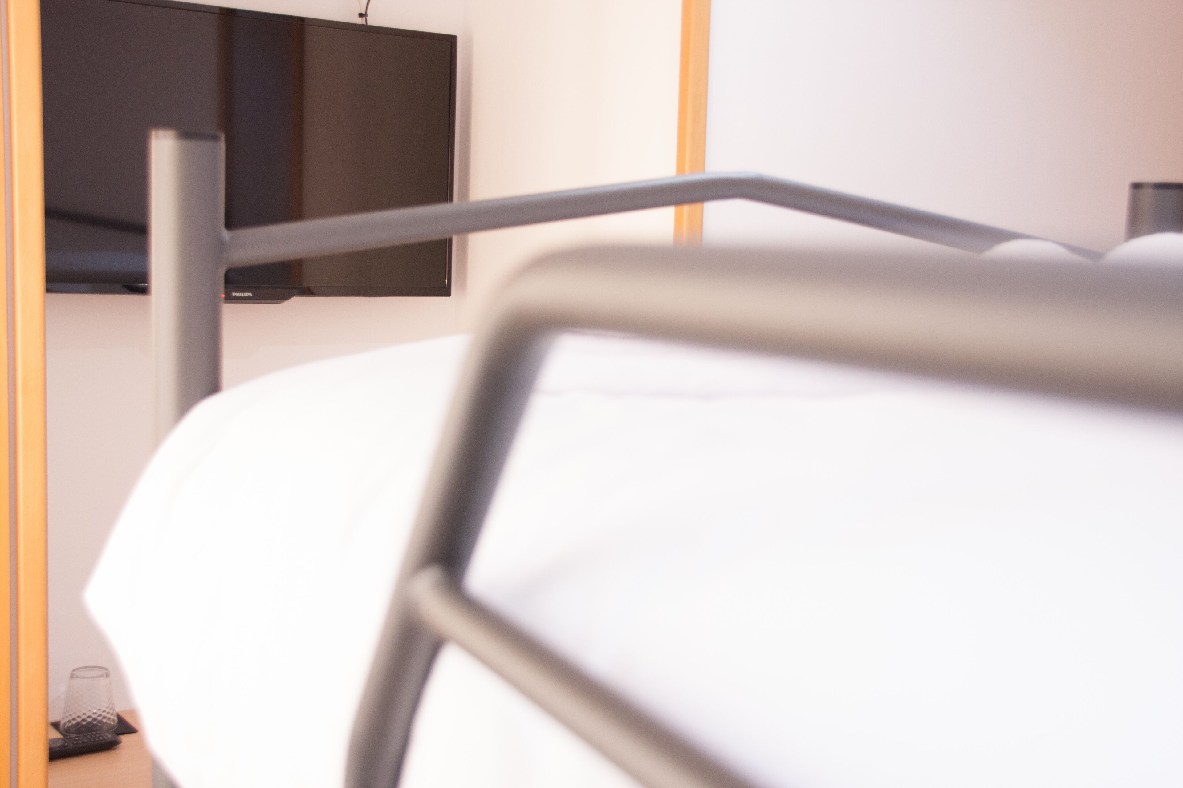 A bed with a metal frame, a television mounted on the wall, and a small table with a glass and a container.