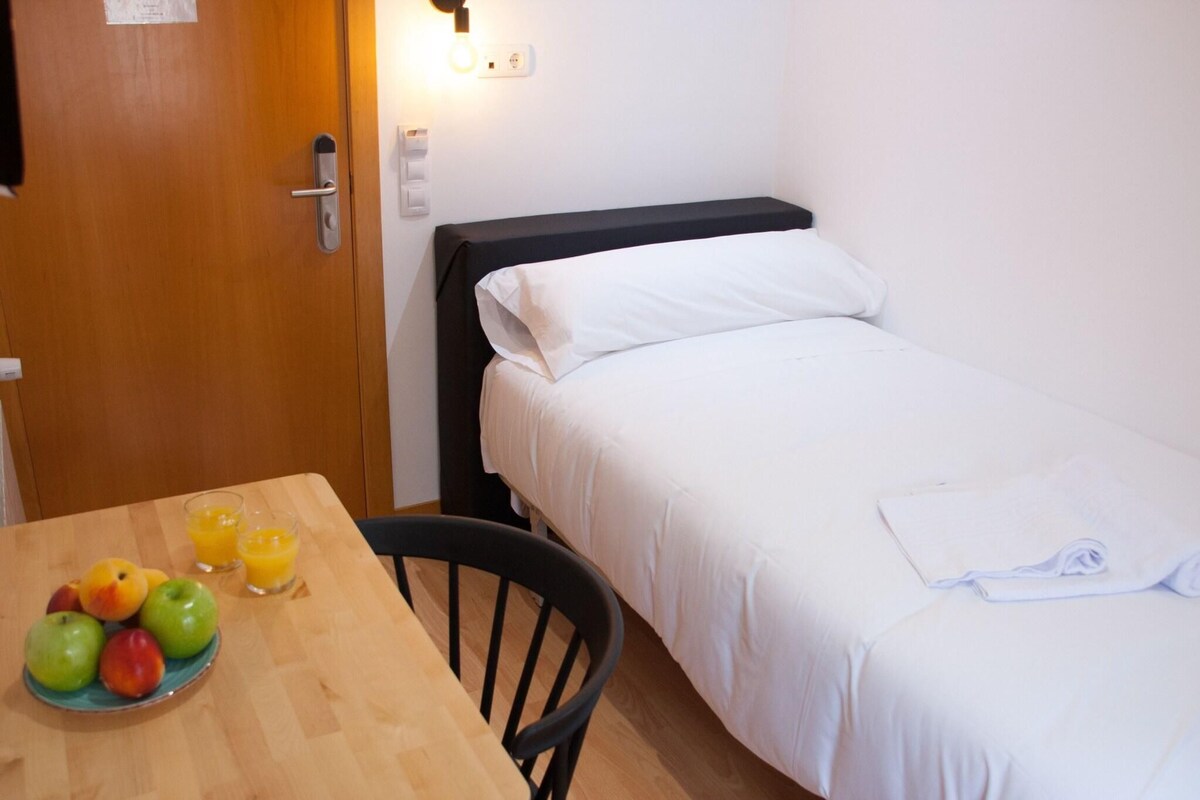 Een kleine, verzorgde slaapkamer met een enkel bed, een nachtkastje met een fruitschaal, en een stoel.