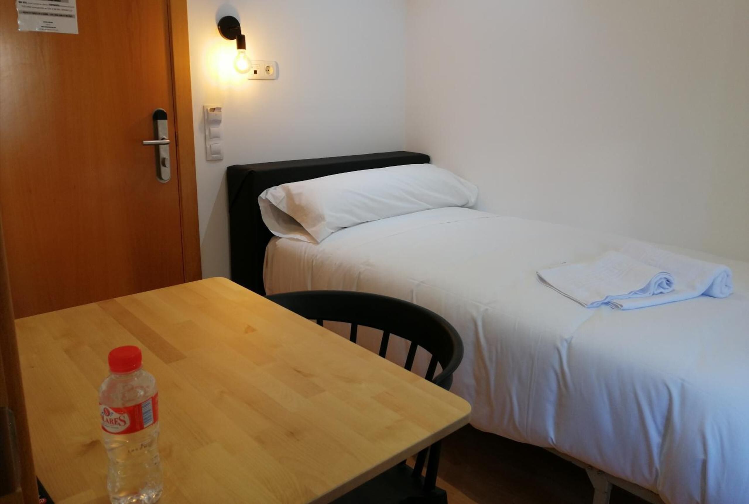 A small, clean room with a wooden table, a single bed with white bedding, a wall-mounted light, and a bottle of water on the table.