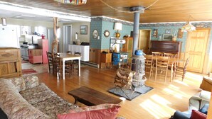 A spacious living room with a wooden floor, a large sofa with a red cushion, a dining area with a wooden table and chairs, a kitchen area with modern appliances, and a decorative chimney.