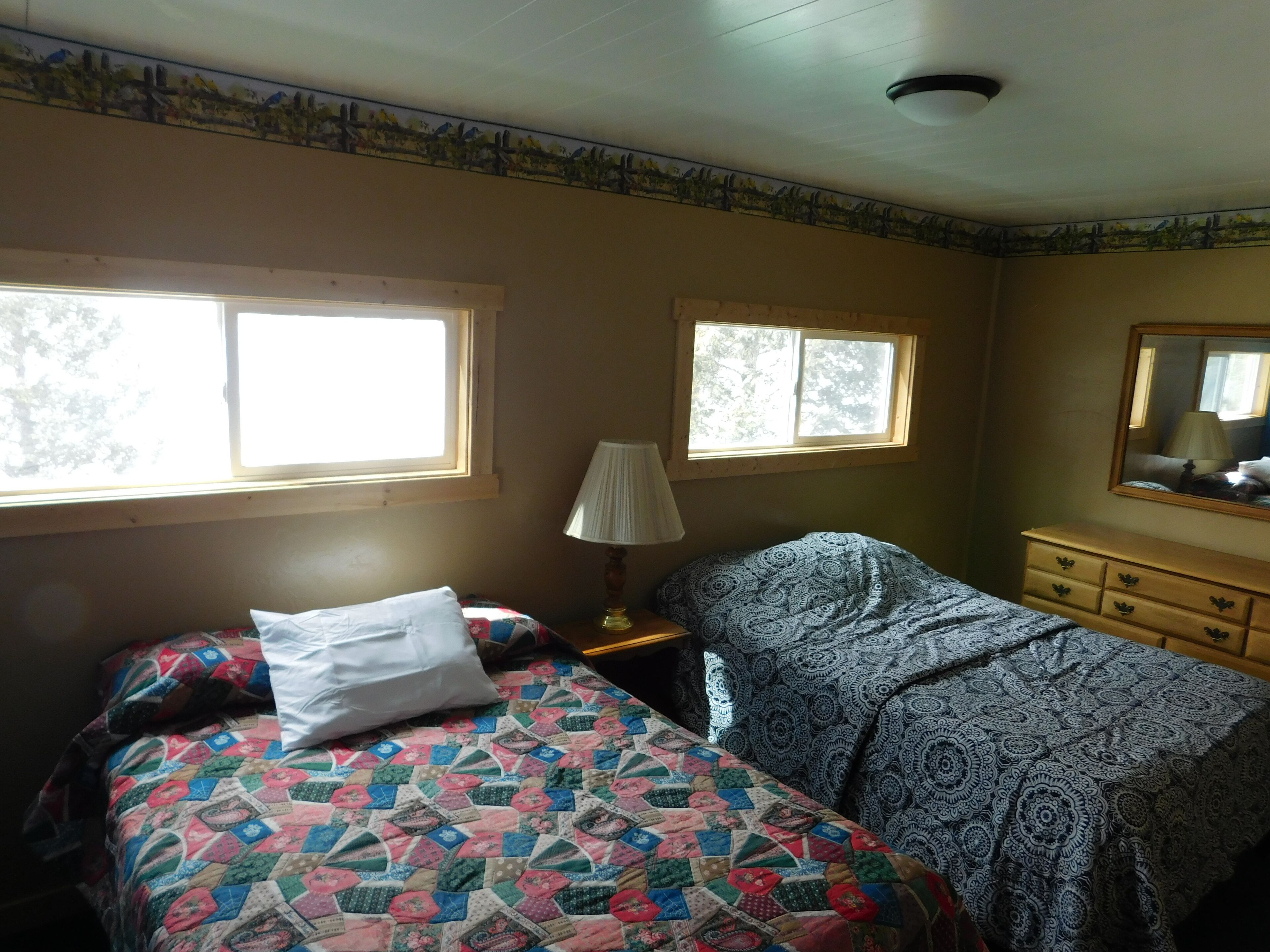 A bedroom with two beds, a nightstand, a lamp, a dresser, and two windows.