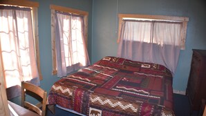 A bedroom with a large bed, a wooden chair, a wooden dresser, and two windows with curtains.