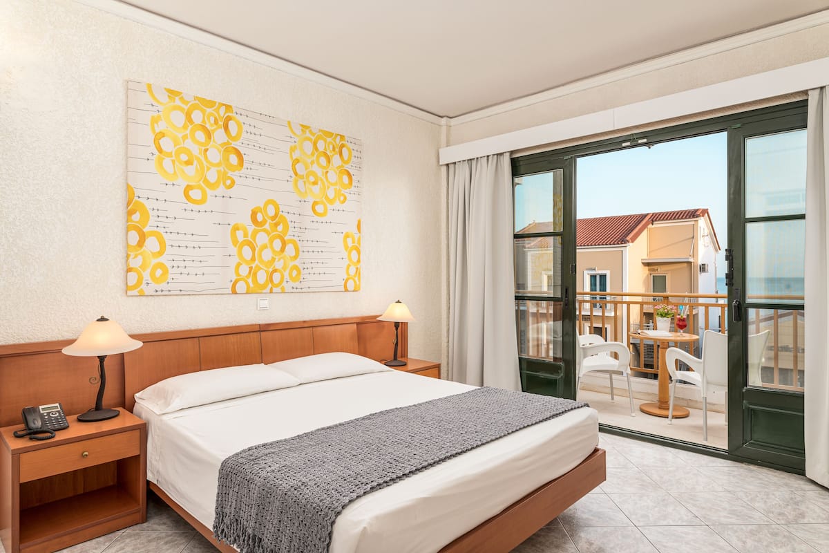 A hotel room with a bed, bedside lamps, a telephone, a large window with a view of the outside, and a balcony with chairs and a table.