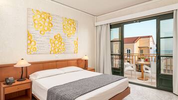 A hotel room with a bed, bedside lamps, a telephone, a large window with a view of the outside, and a balcony with chairs and a table.