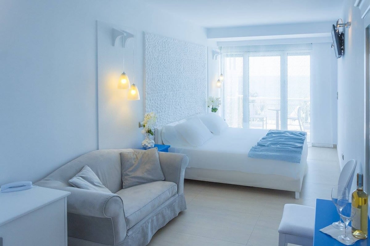 A modern hotel room with a bed, a sofa, a desk, and a balcony with a view.