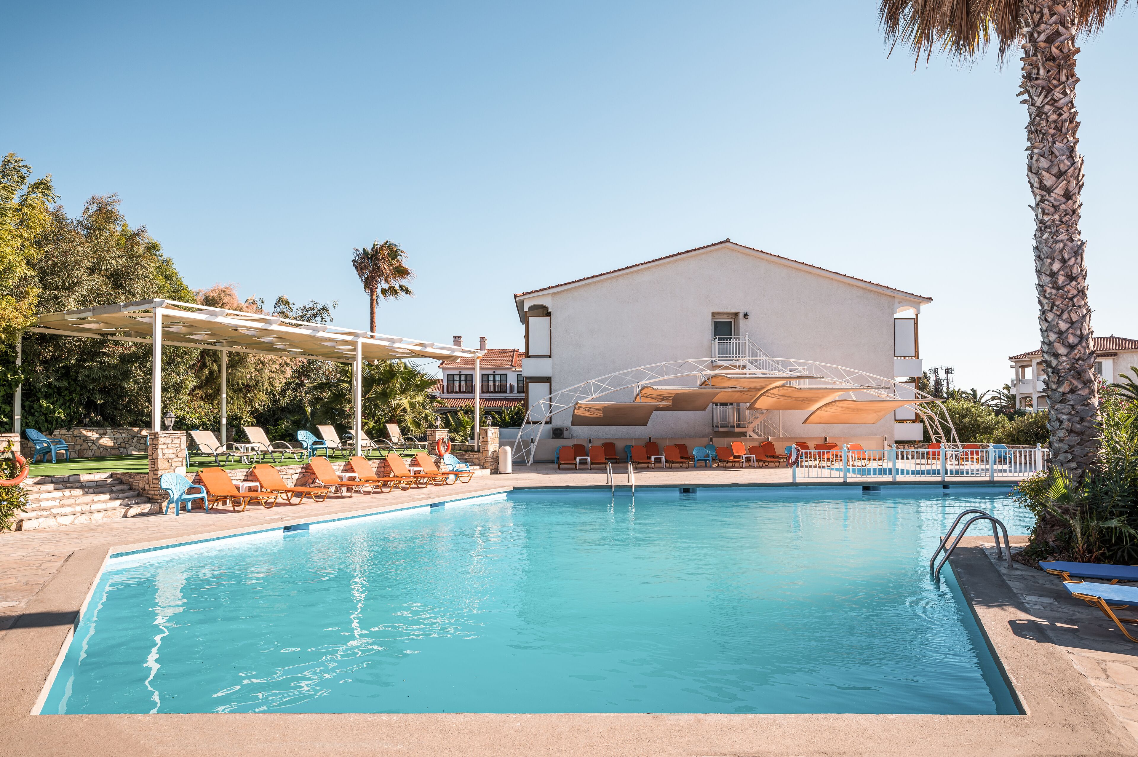 A swimming pool with lounge chairs and a covered seating area, surrounded by palm trees and a building with a white facade.