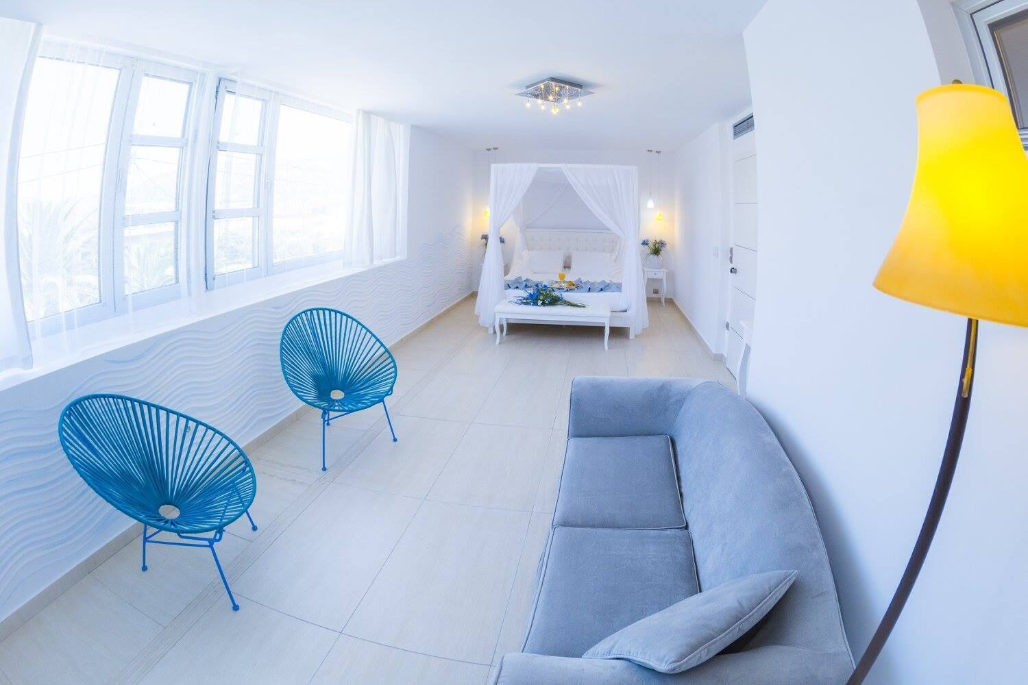 Un salon lumineux et moderne, avec un canapé gris, deux fauteuils bleus, un lit à baldaquin et une lampe de sol jaune.