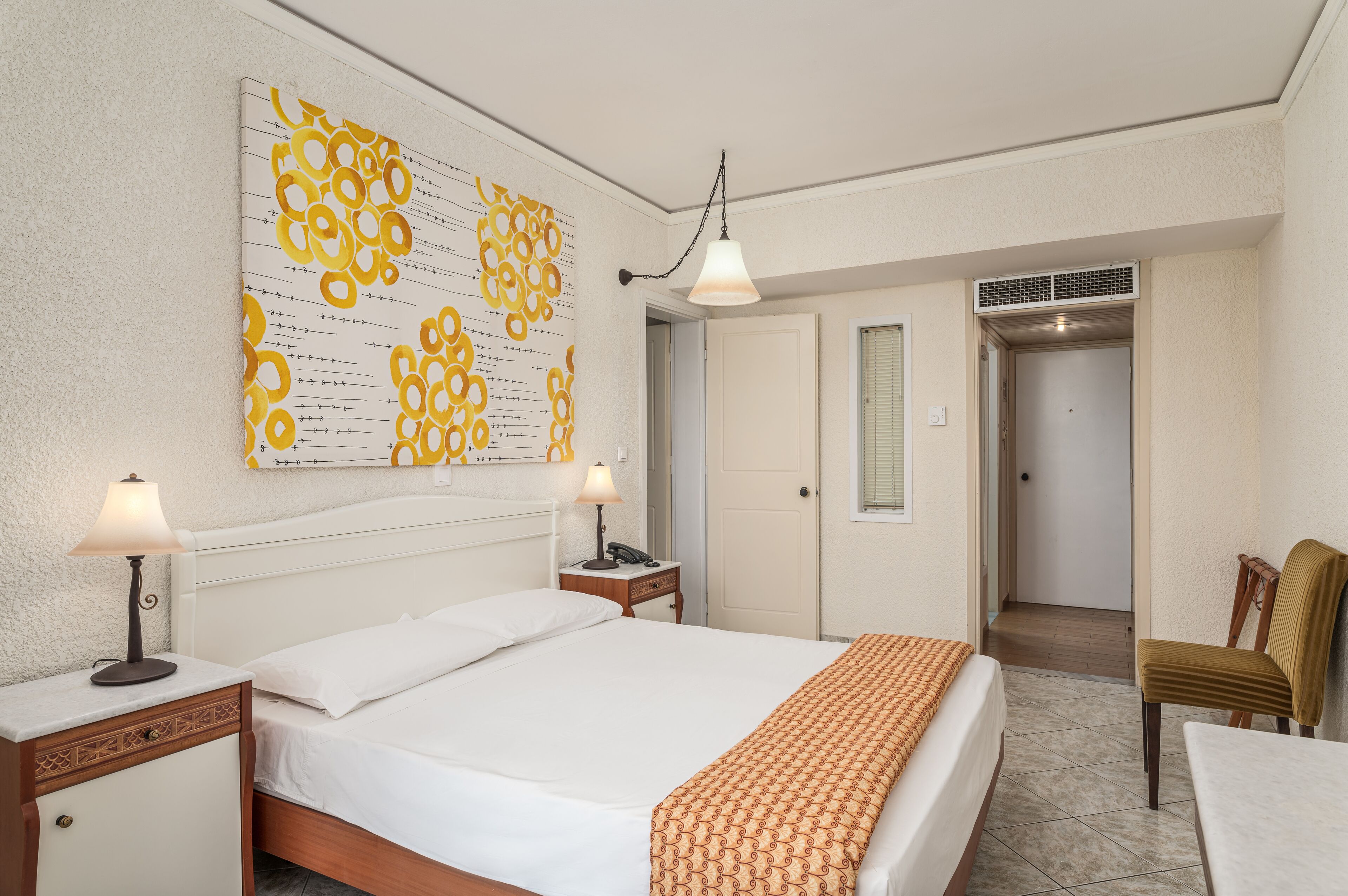 Habitación de hotel con una cama grande, dos mesitas de noche con lámparas, un tapiz decorativo en la pared y una puerta que conduce a otra habitación.
