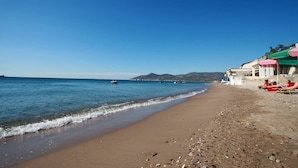A beach with clear blue water, a sandy shore, and a few boats. There are buildings with colorful roofs in the background.