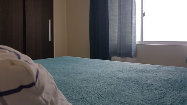 A bedroom with a bed, a pillow, a window with curtains, and an air conditioning unit on the wall. - Apartment between the airport and the beach. (Boa Viagem)