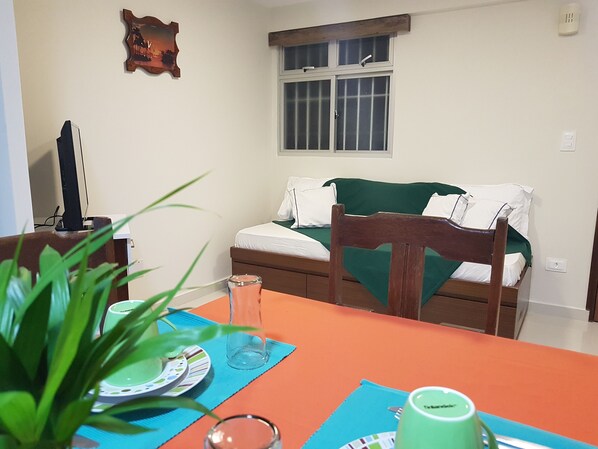 A room with a bed, a table with a plant, a green mug, and a window with blinds.