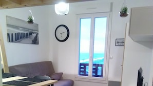 En kompakt bolig med træloft, et spisebord med stole, en sofa, et lille hvidt skab og et vindue med havudsigt.