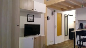 En kompakt opholdsstue med et fjernsyn på et hvidt skab, et lille spisebord med stole, og en dør med en gul, struktureret panel.
