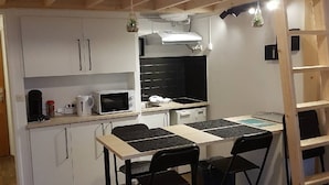 Et kompakt køkken med hvide skabe, en sort bordplade, en mikroovn og en gasblus. Der er to sorte stole og en træstige.