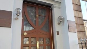 Une porte en bois brun, dotée de panneaux de verre et d’une poignée dorée, flanquée de colonnes blanches et de deux appliques murales.