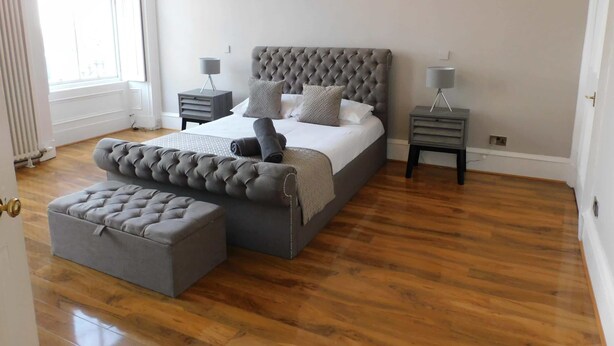 Une chambre à coucher avec un grand lit, deux tables de chevet, un banc recouvert d’un tissu gris et un radiateur.
