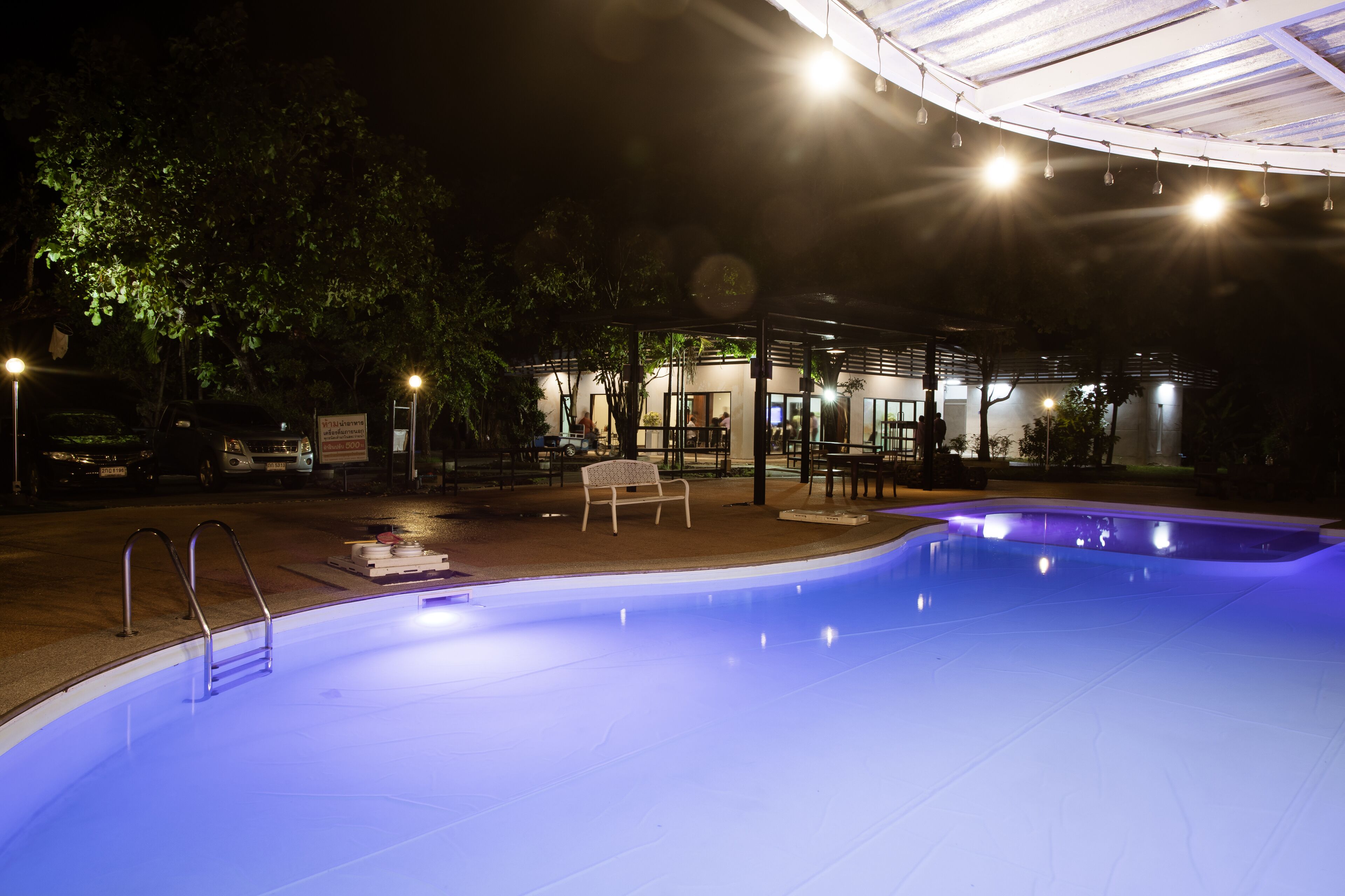 Una piscina con escalera, rodeada por una zona de estar cubierta con mesas y sillas, y un edificio con ventanas amplias al fondo.