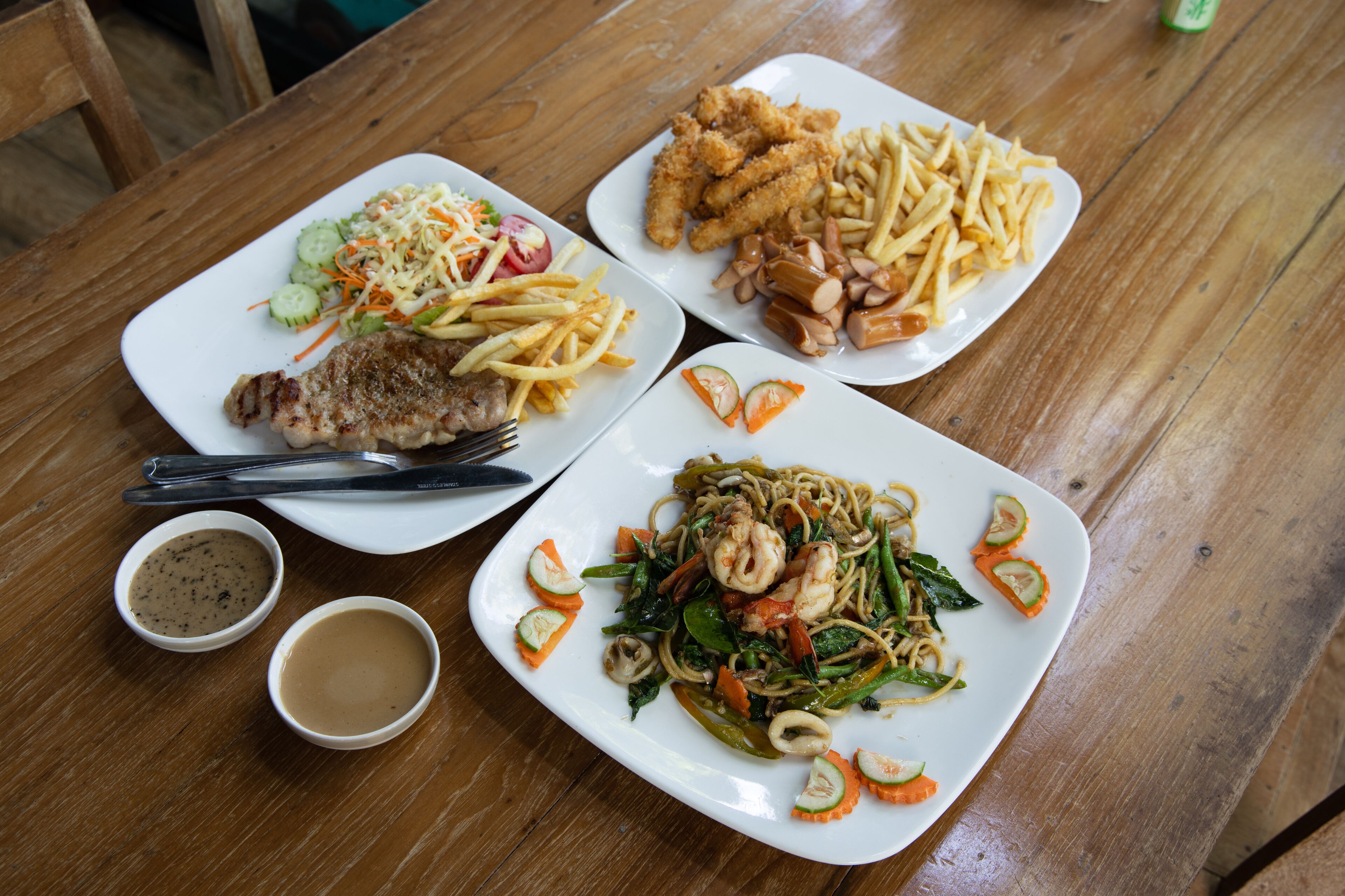A wooden table with three square plates of food, including a steak, fries, and stir-fried vegetables.