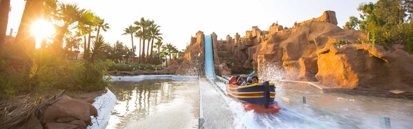 A water ride with a blue and yellow boat, surrounded by rocky cliffs and palm trees.