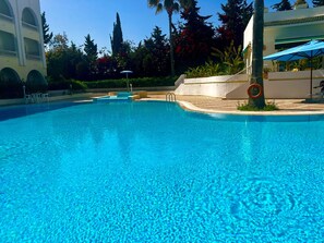 A clear blue swimming pool with a palm tree and a blue umbrella nearby.