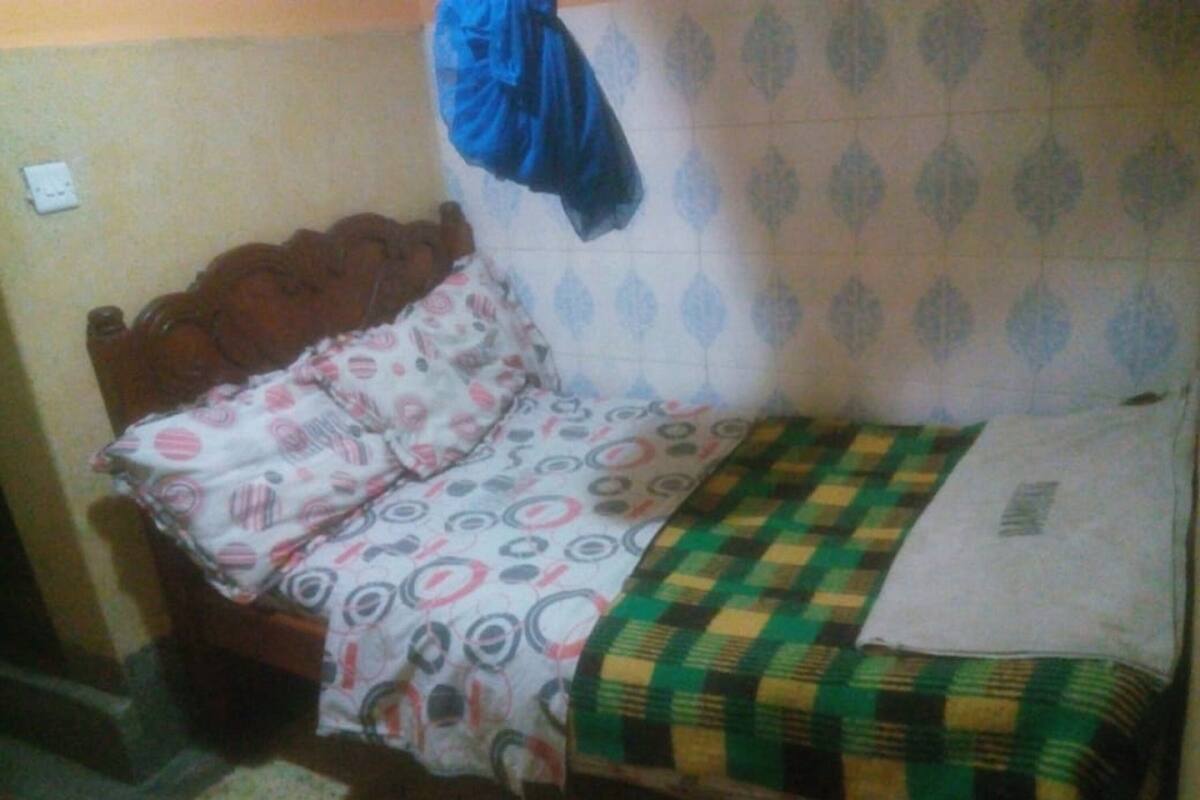 Quarto pequeno e simples com cama de madeira, roupa de cama estampada e uma parede lisa com tomada.