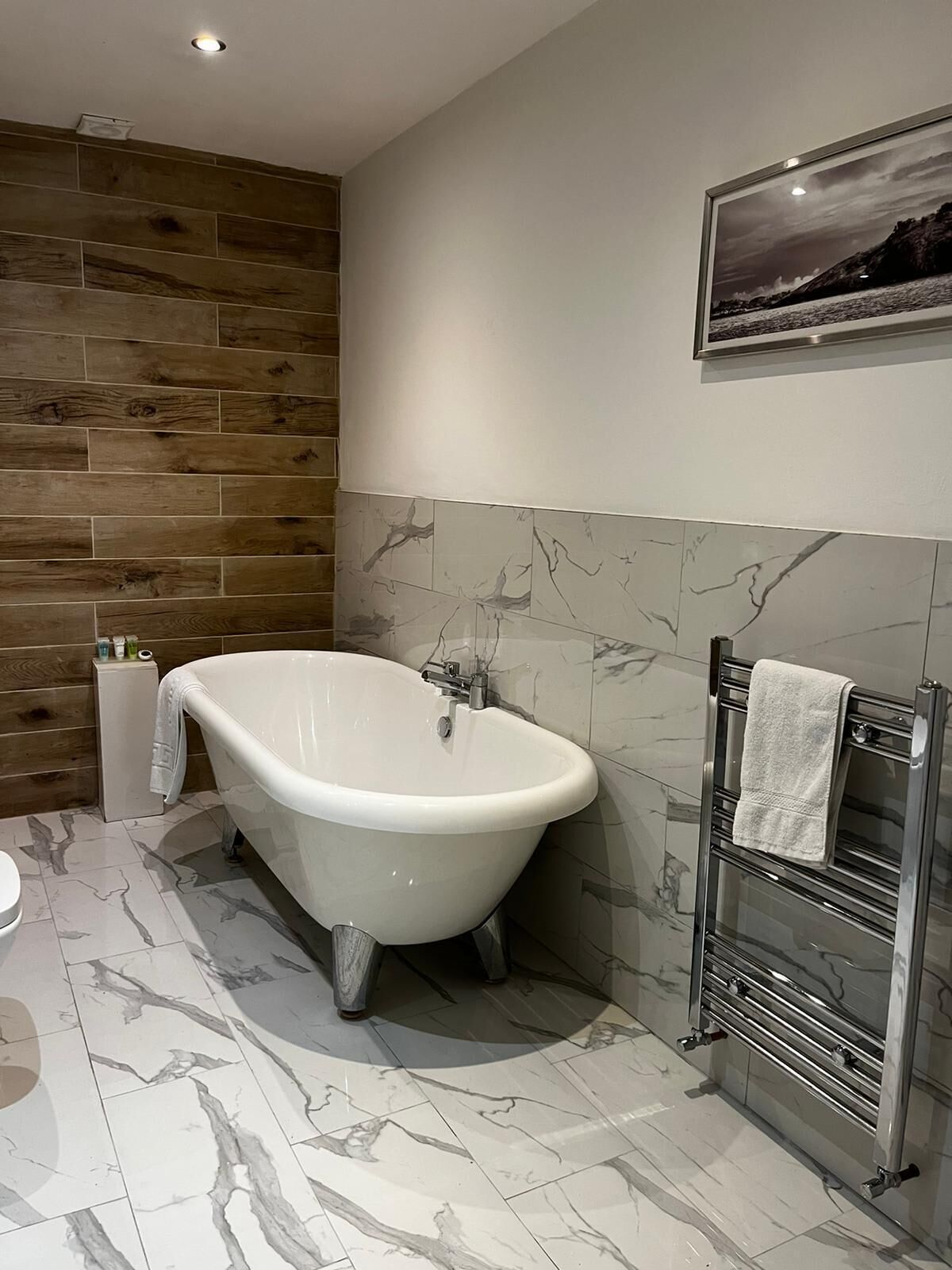 Une salle de bain moderne avec une baignoire sur pieds, un revêtement de sol en marbre, un mur d’accent en bois et un porte-serviettes.