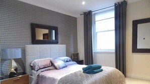 Une chambre à coucher avec un lit, une table de chevet, une lampe, un miroir et une fenêtre avec des rideaux.