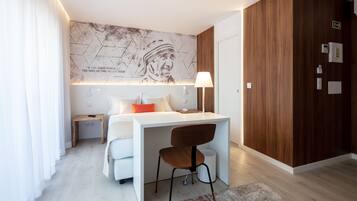 A modern bedroom with a white bed, a desk, and a chair. There is a map on the wall, a lamp, and a small nightstand.