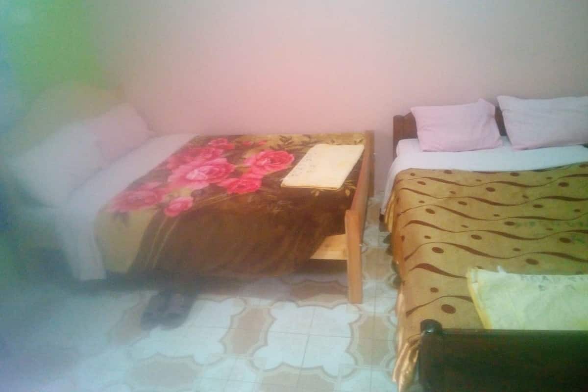 A room with two beds, one with a floral bedspread and the other with a patterned bedspread. There is a small table with a floral table runner between the beds.