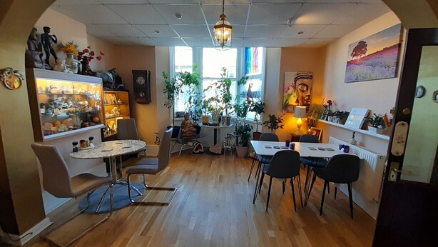 A cozy interior with wooden flooring, a central round table, and chairs, surrounded by shelves with various items and a large window with plants. - Starlight Guest House (Blackpool)