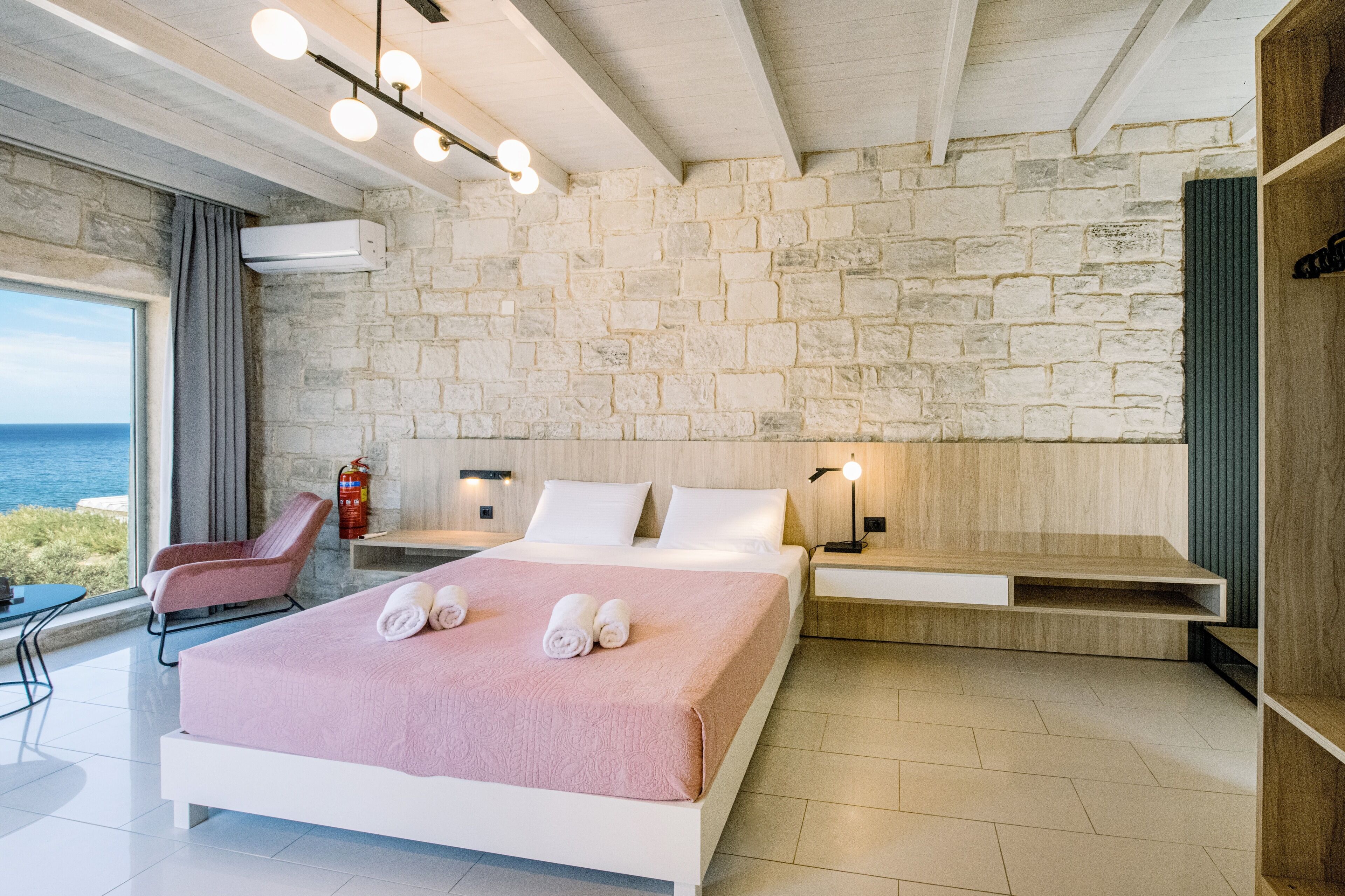 A modern hotel room with a stone wall, a bed with a pink blanket, a bedside table, a chair, and a view of the sea.
