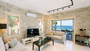 Sala de estar con paredes de piedra, un televisor de pantalla plana, un sofá, una mesa de centro y vistas al mar a través de amplias ventanas.