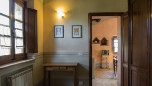 A room with a wooden table, a radiator, and a window with curtains. There are framed pictures on the wall and a door leading to another room. - NOBLE FARM HOLIDAY APARTMENT (Montepulciano Siena)